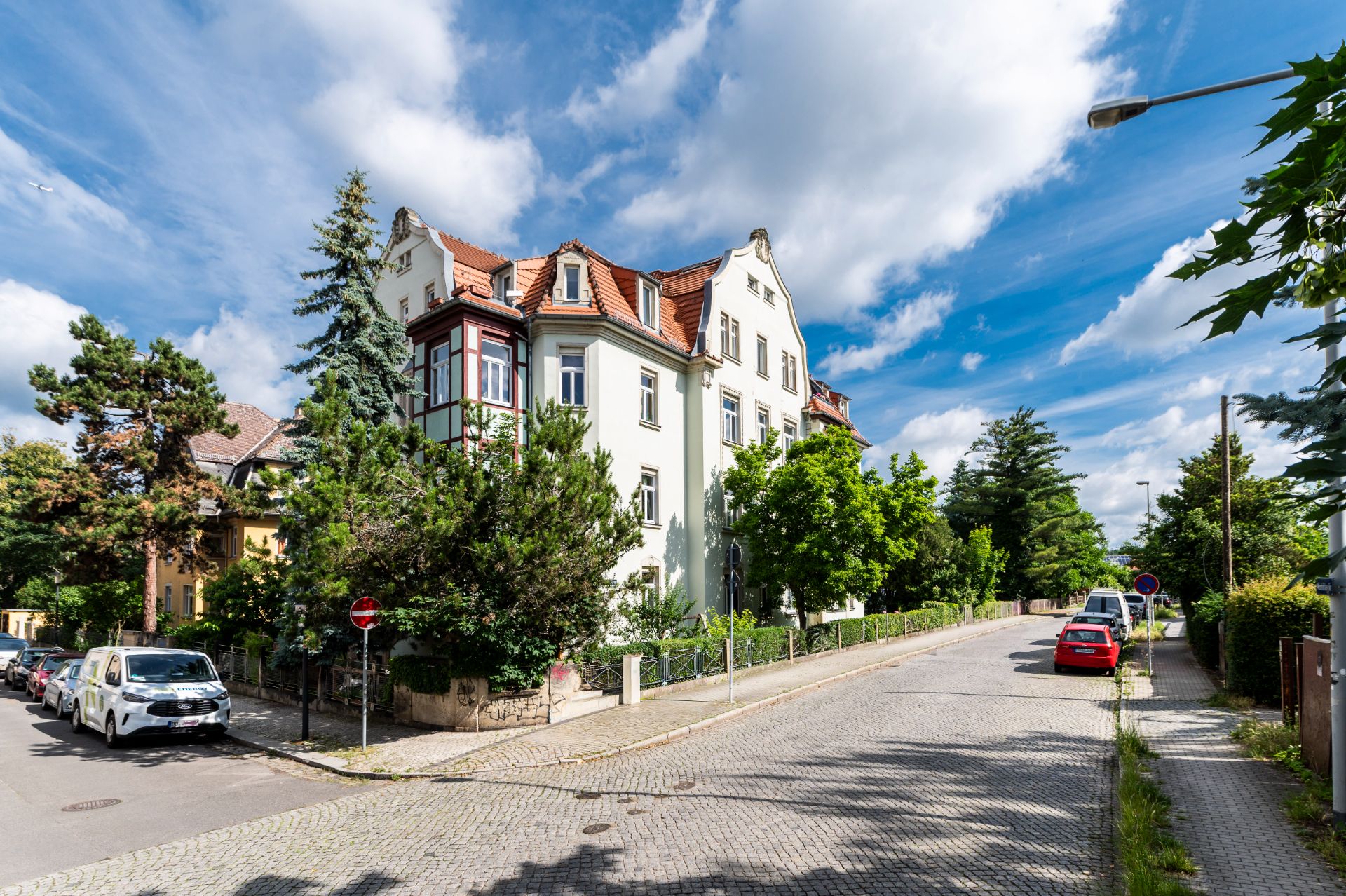 Eckhaus mit rotem Dach an einer ruhigen, baumbestandenen Wohnstraße bei blauem Himmel.