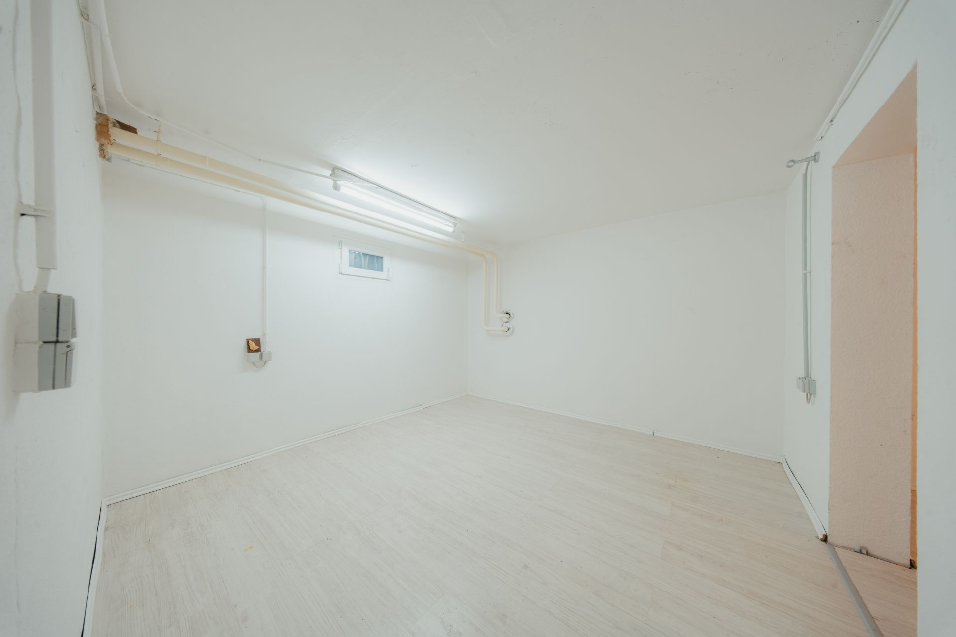 Leerer, weiß gestrichener Kellerraum mit hellem Boden und kleinem Fenster an der Wand.