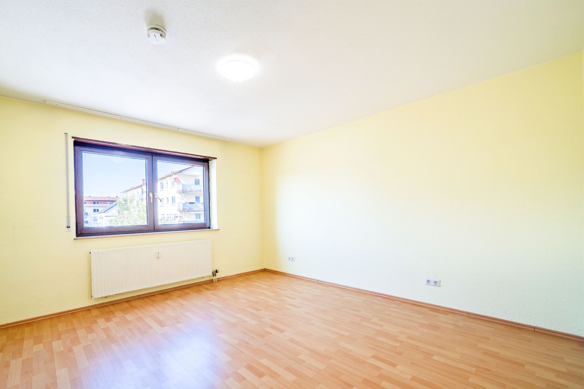 Heller leerer Raum mit Holzfußboden und großem Fenster, gelbe Wände, Heizkörper unter dem Fenster.