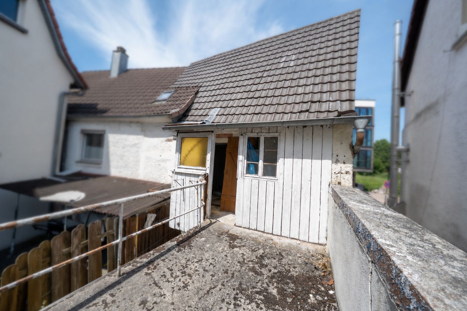 Kleines weißes Gebäude mit schrägem Dach und offen stehender Tür auf einer Terrasse.