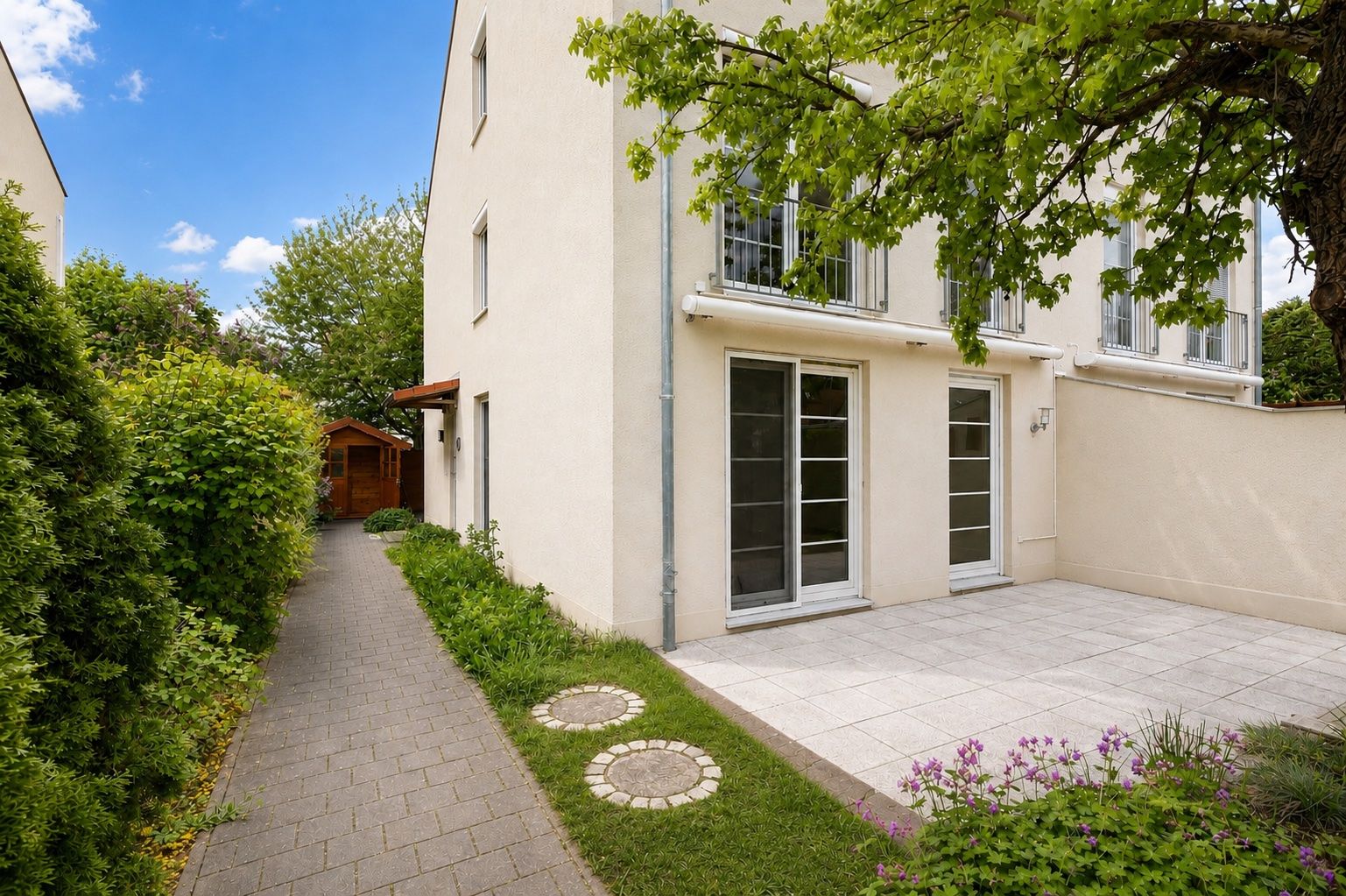 Terrasse mit Fliesen neben einem hellen Wohnhaus, davor Gartenweg und Baum.