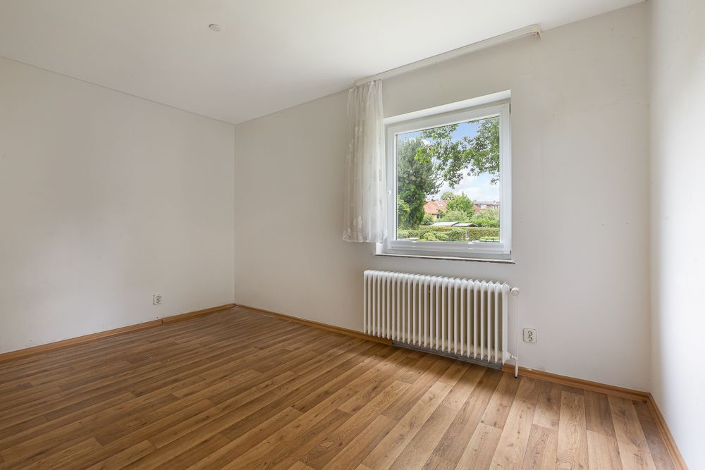 Leeres Zimmer mit Holzfußboden, weißer Wand, Fenster mit Vorhang und Heizkörper unter dem Fenster.