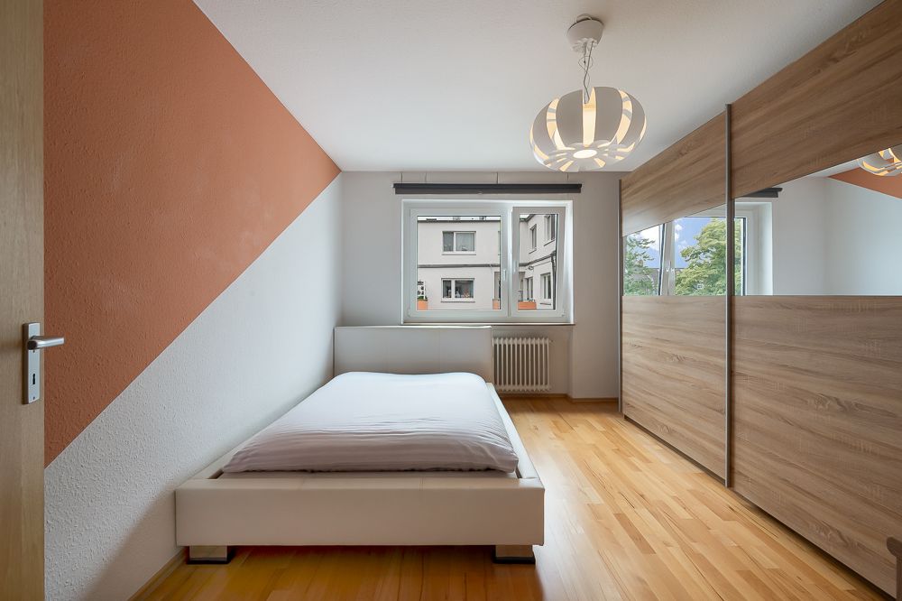 Schlafzimmer mit Einzelbett, großem Kleiderschrank und Fenster, Holzboden und moderner Deckenlampe.
