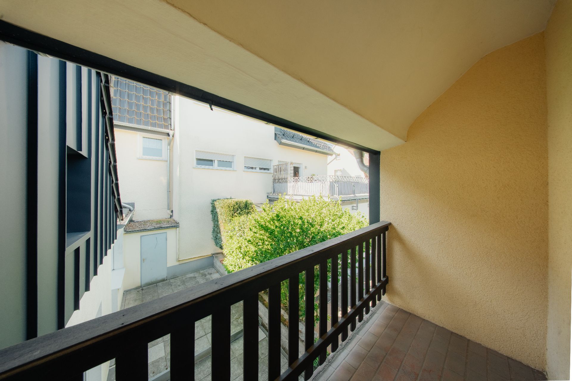Überdachter Balkon mit Holzboden und schwarzem Geländer, Blick auf Nachbarhäuser und Garten.
