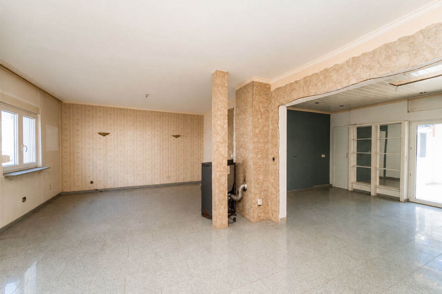 Geräumiges, leeres Wohnzimmer mit großen Fenstern, offenem Durchgang und heller Fliesenboden.