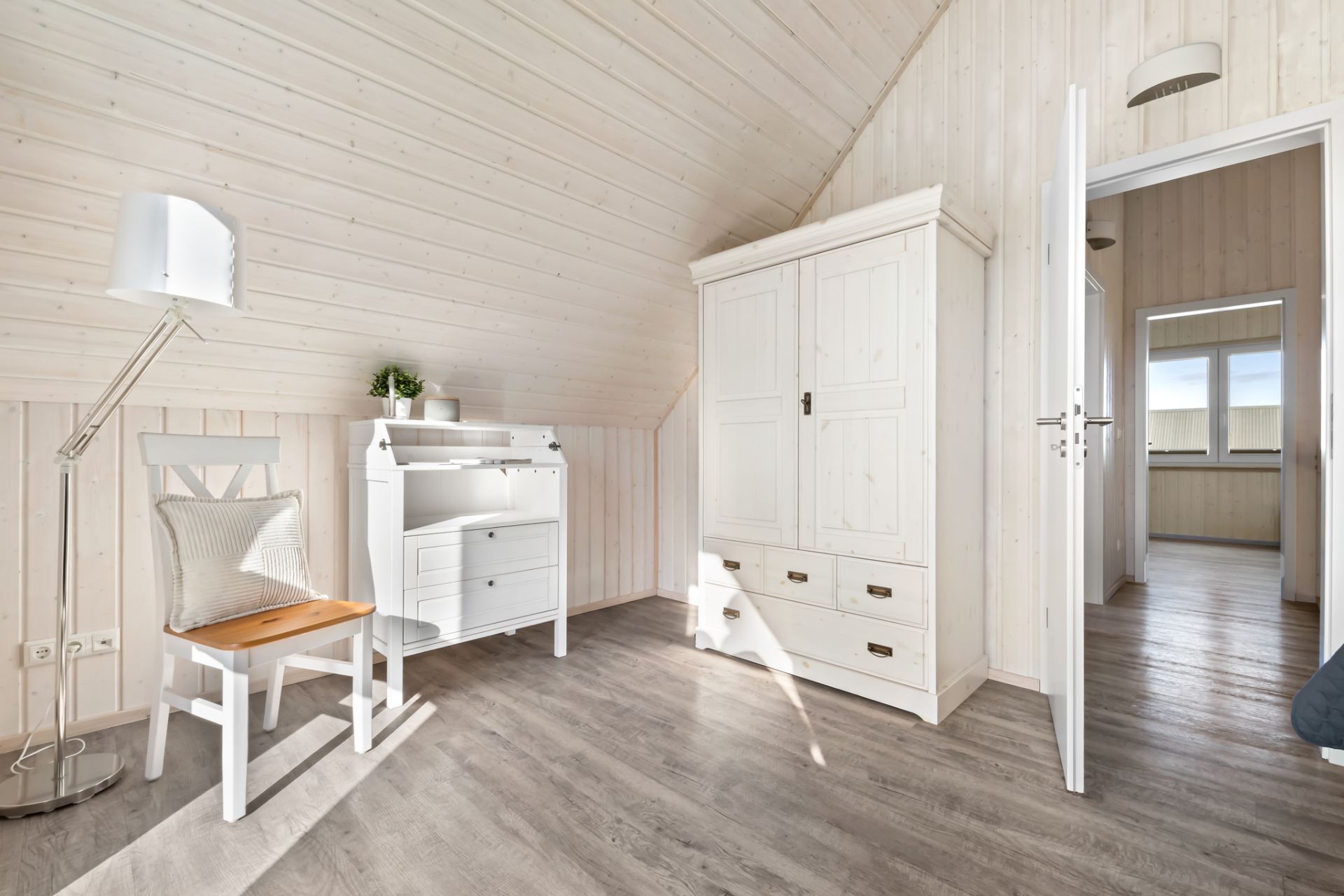 Helles Zimmer mit Schreibtisch, Stuhl, Stehlampe und weißem Kleiderschrank auf Holzfußboden.