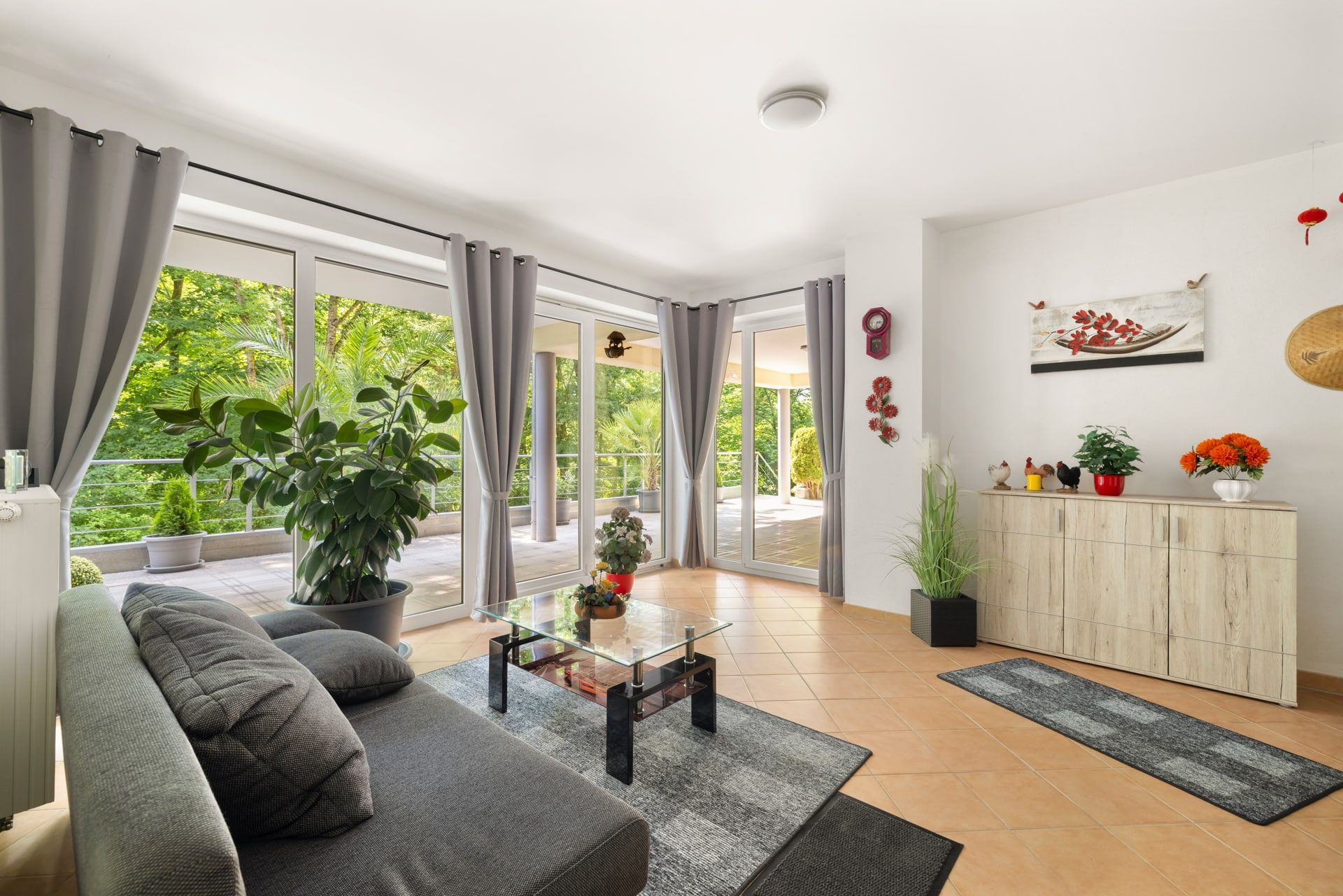 Helles Wohnzimmer mit grauem Sofa, Glastisch, Pflanzen und großen Fenstern zur Terrasse.
