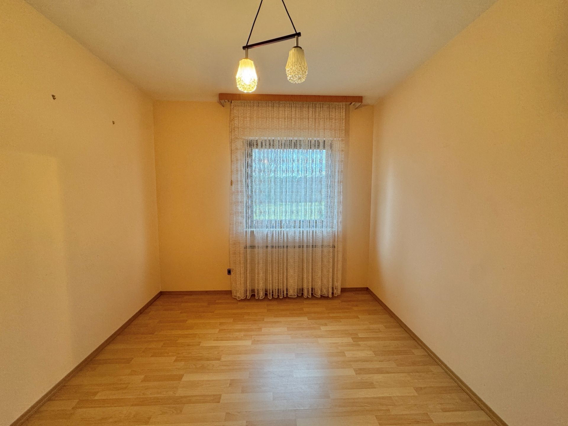 Leerer Raum mit hellem Holzboden, Fenster mit Gardine und zwei Deckenlampen.