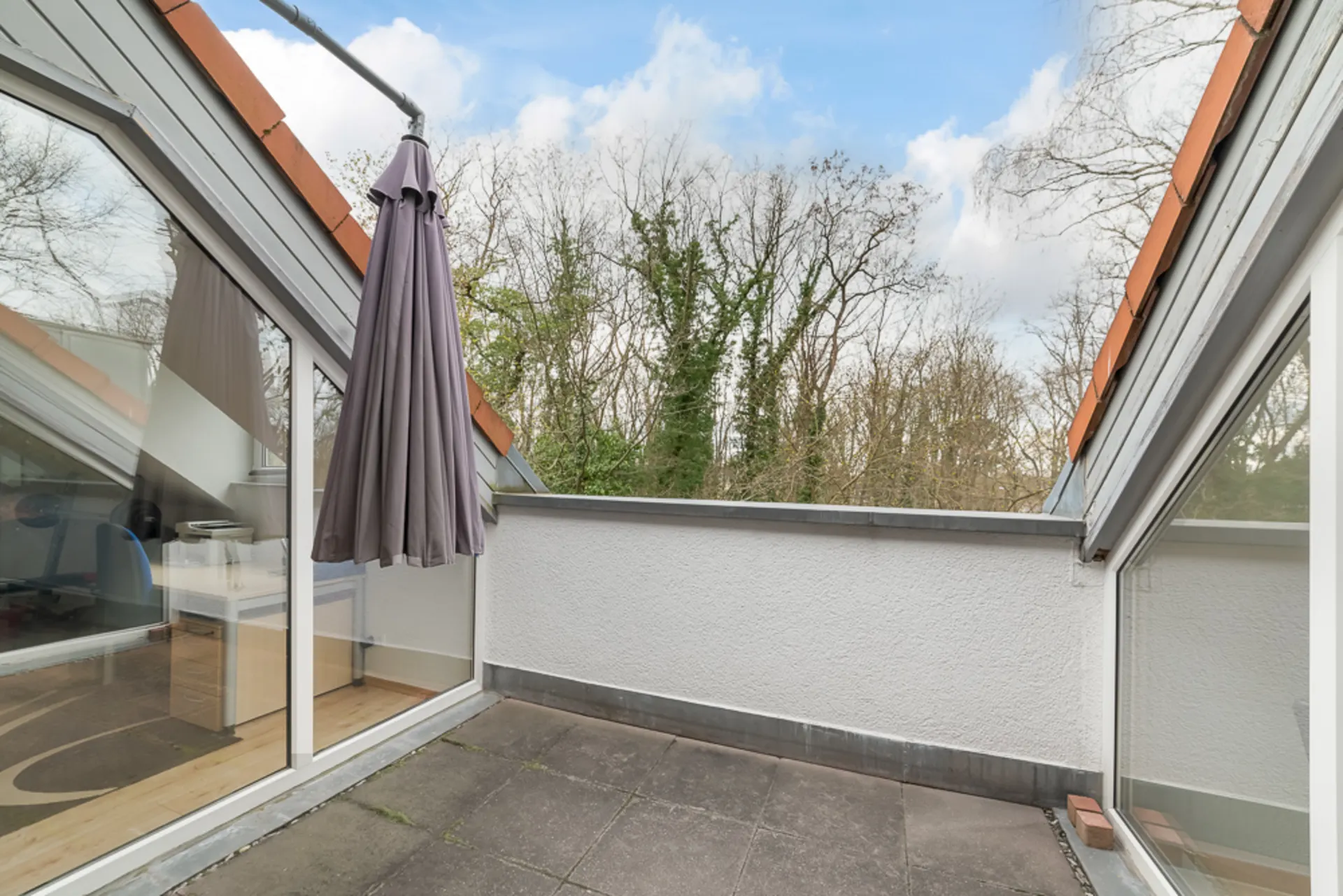 Dachterrasse mit geschlossenem Sonnenschirm und Blick auf Bäume, umgeben von Glas und weißen Wänden.