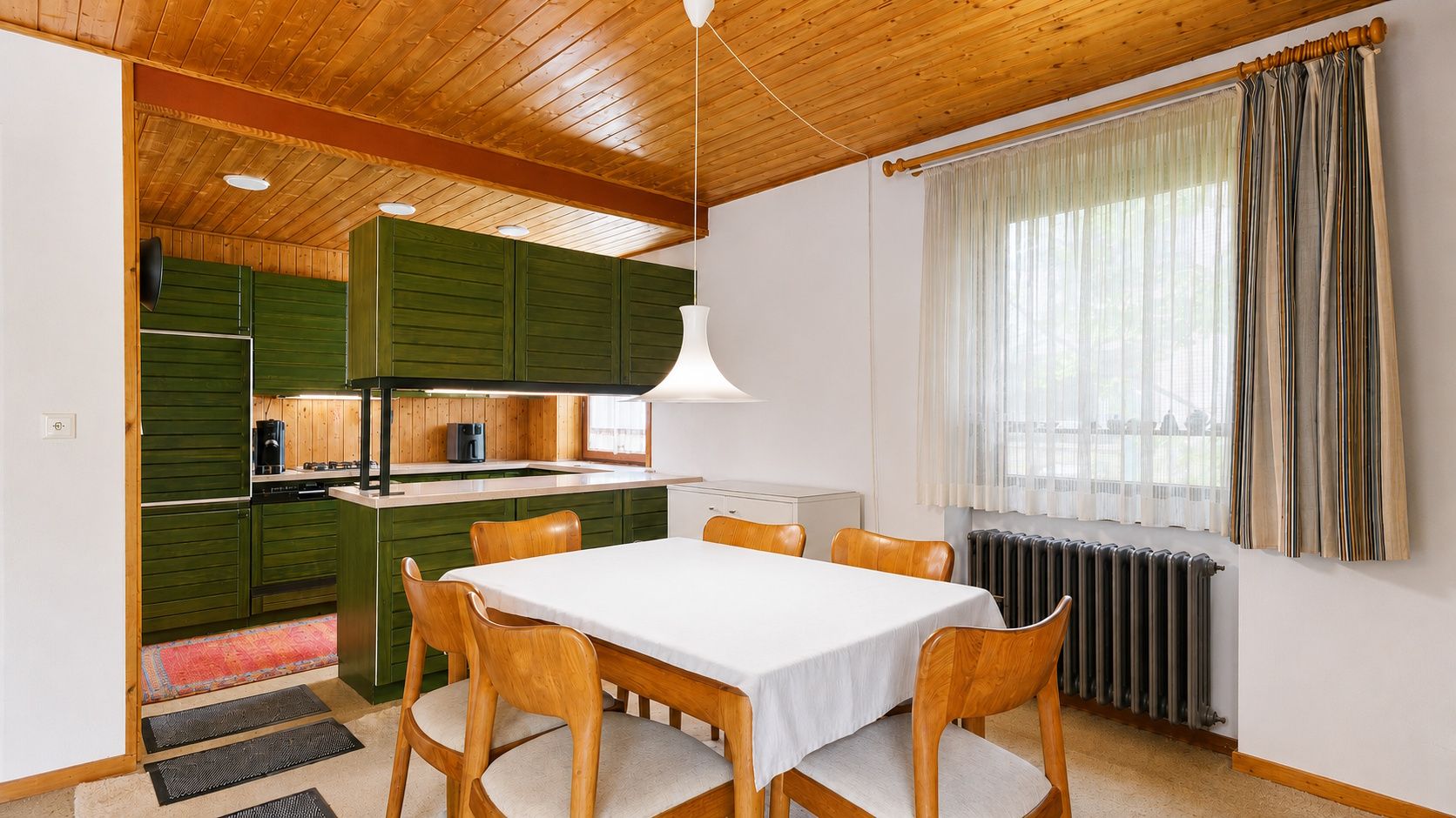 Essbereich mit Holztisch und sechs Stühlen, daneben grüne Einbauküche und Fenster mit Vorhang.