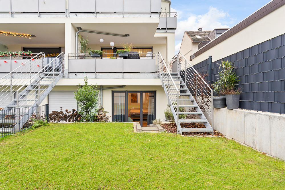 Grüner Garten mit Terrasse, zwei Treppen und Balkonen an einem modernen Wohnhaus.