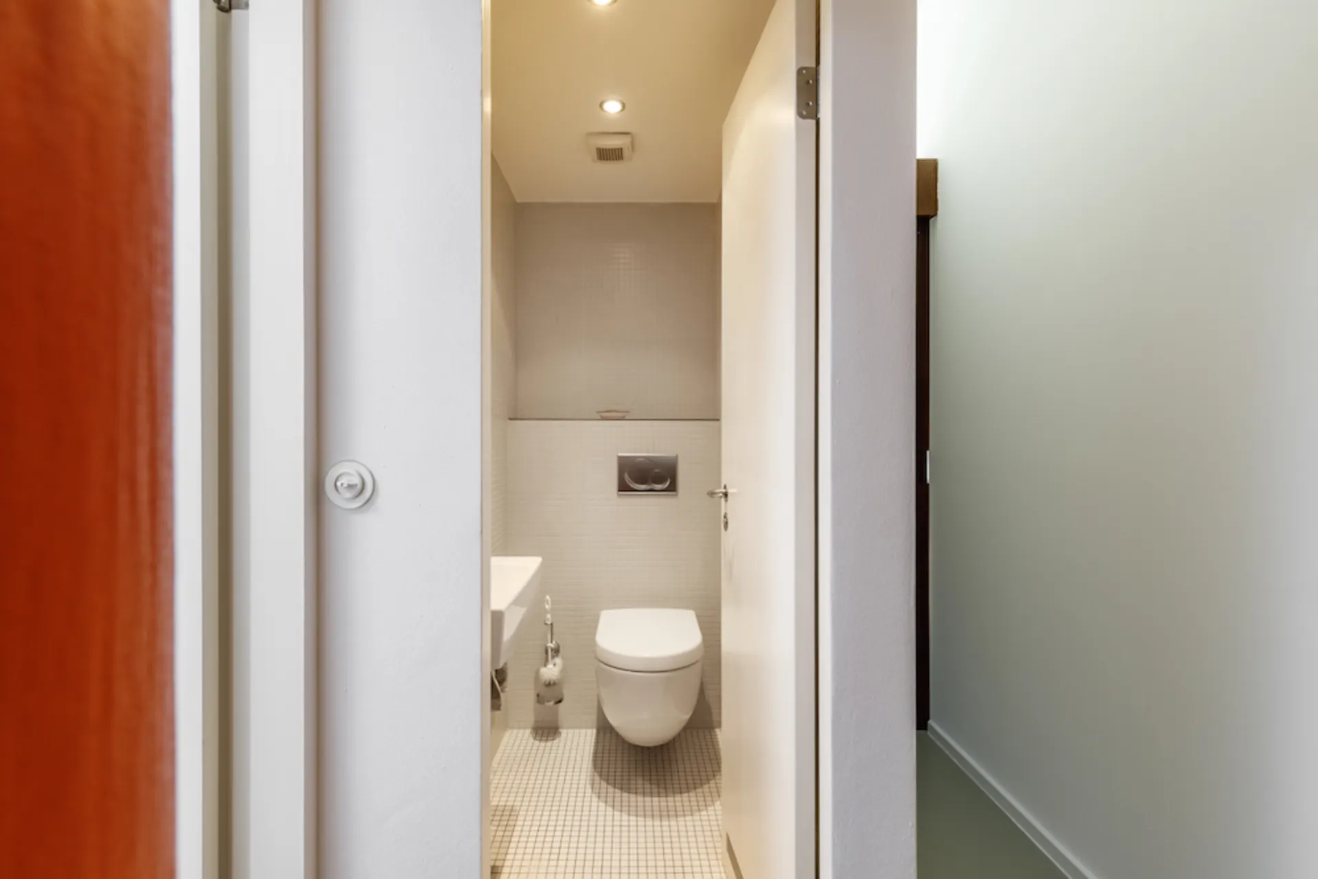 Kleine Toilette mit wandhängendem WC, kleinem Waschbecken und heller Beleuchtung.