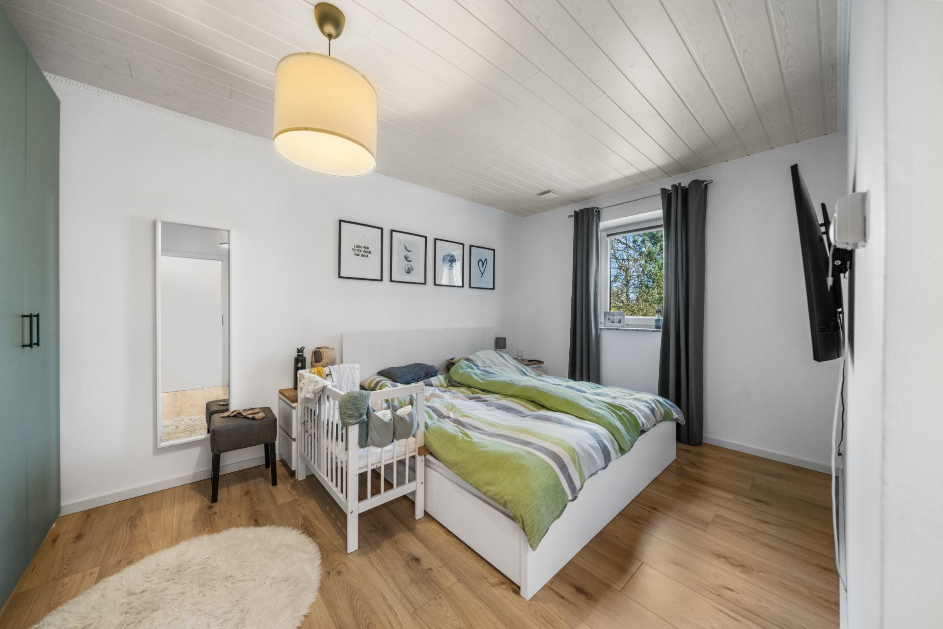 Schlafzimmer mit Doppelbett, Kinderbett, Spiegel und Fenster mit Vorhängen.
