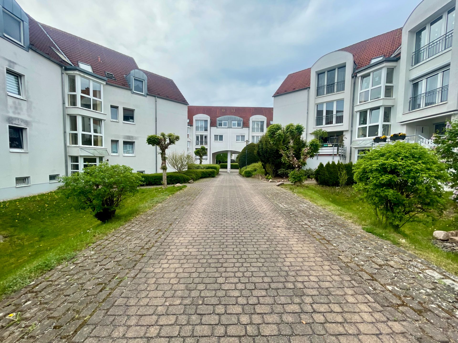 Gepflasterter Weg zwischen weißen Wohngebäuden mit roten Dächern und grünen Büschen.