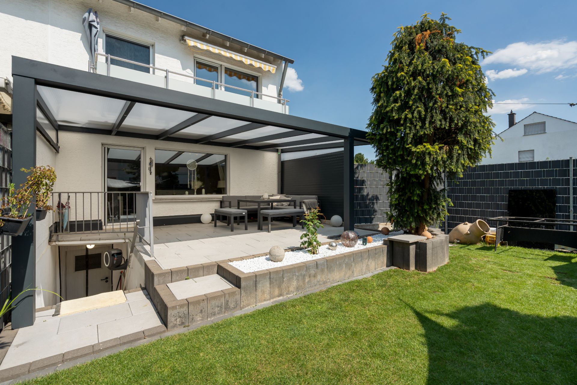 Überdachte Terrasse mit Essbereich, Garten und Baum vor einem Wohnhaus.