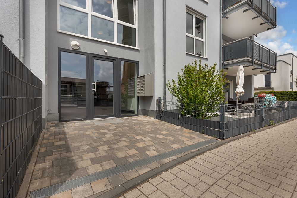 Gepflasterte Terrasse mit Zaun und großen Fenstern an einem modernen Wohngebäude.