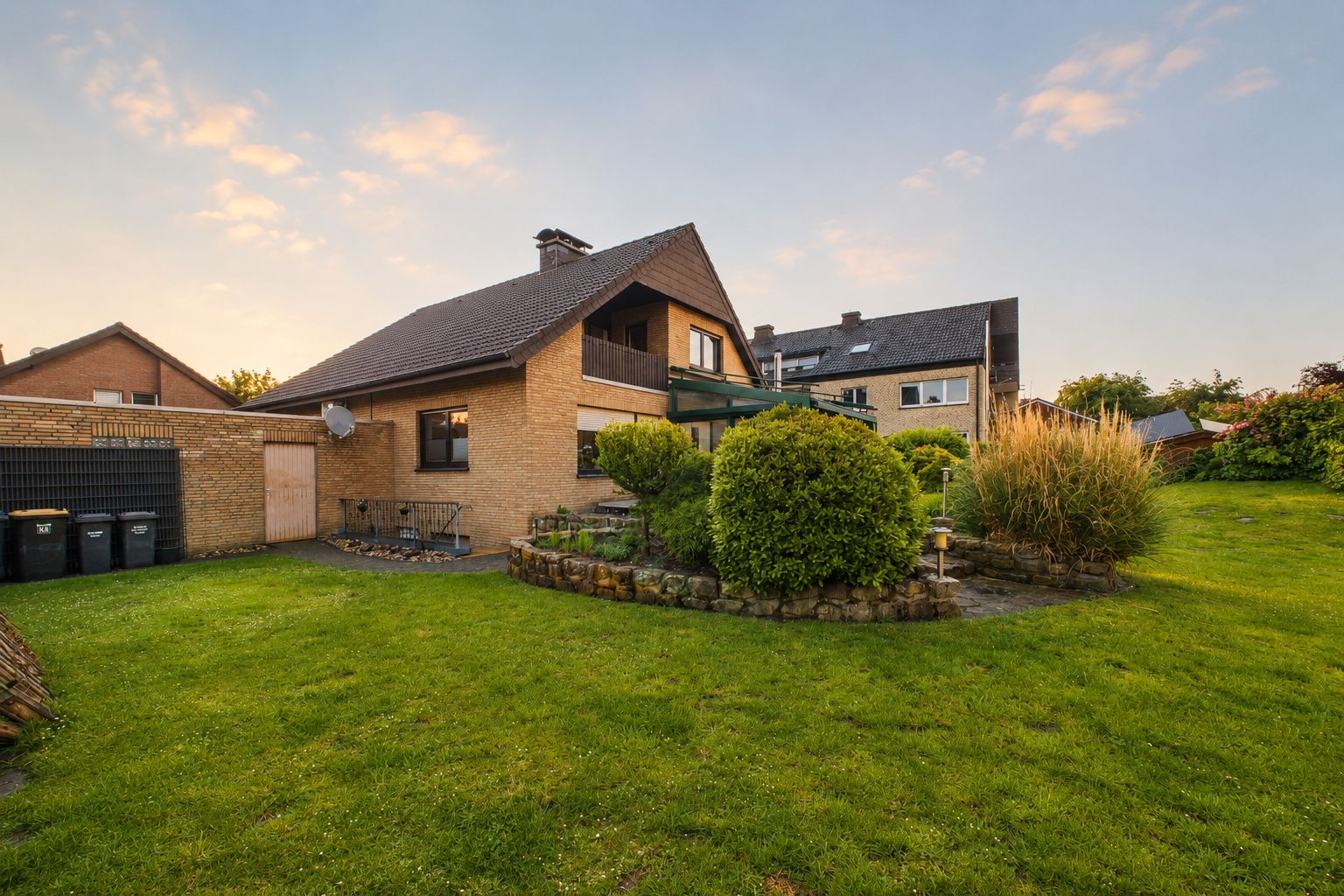Einfamilienhaus mit Garten, Terrasse und Sträuchern bei Sonnenuntergang.
