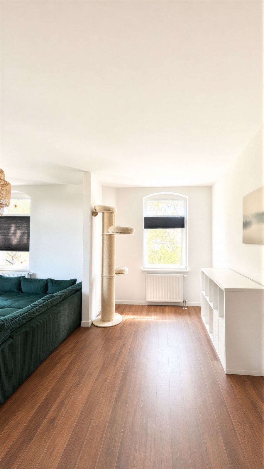 Helles Wohnzimmer mit grünem Sofa, Kratzbaum, Regal und großem Fenster mit schwarzem Rollo.