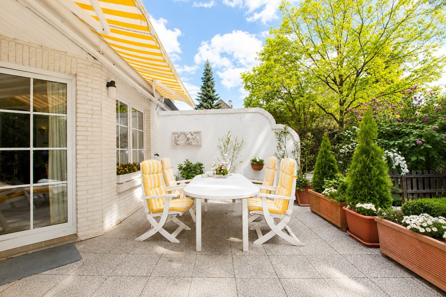 Helle Terrasse mit weißem Tisch, vier gelb-weißen Stühlen und Pflanzen in Blumentöpfen.