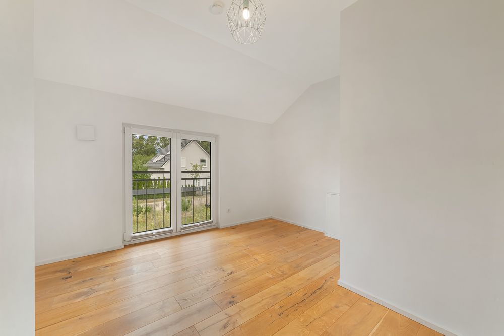 Leeres Zimmer mit Holzfußboden, weißen Wänden und bodentiefen Fenstern mit Blick nach draußen.