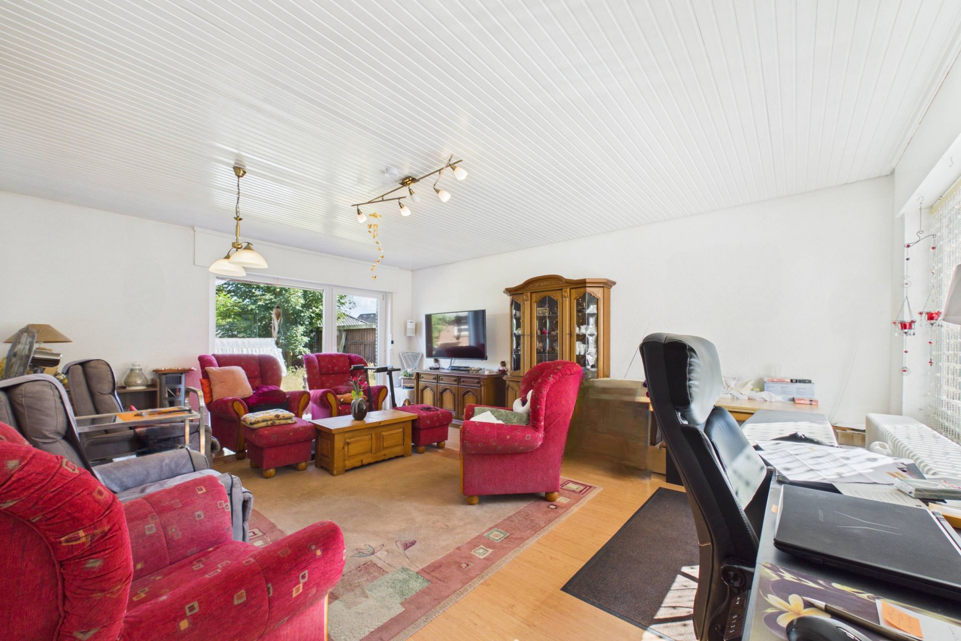 Wohnzimmer mit roten Sesseln, Holzmöbeln, Fernseher und großem Fenster, heller Raum.