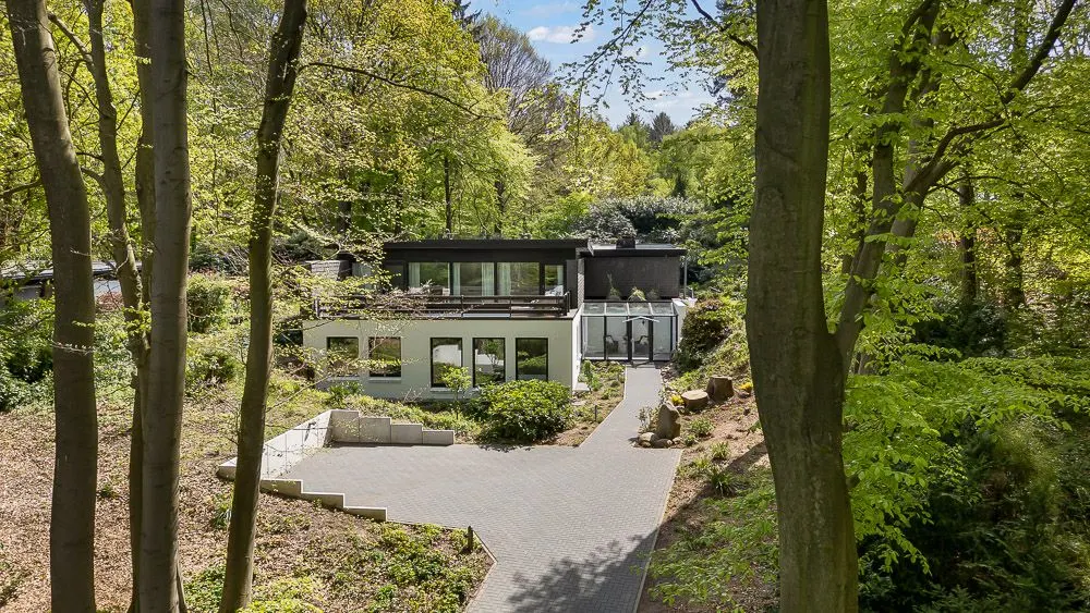 Modernes Haus mit zwei Etagen und großer Fensterfront im Wald, Zufahrt und Garten sichtbar.