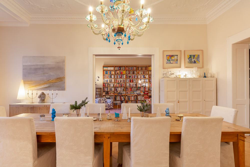 Esszimmer mit großem Holztisch, weißen Stühlen, Kronleuchter und Blick in ein Zimmer mit Bücherregal.