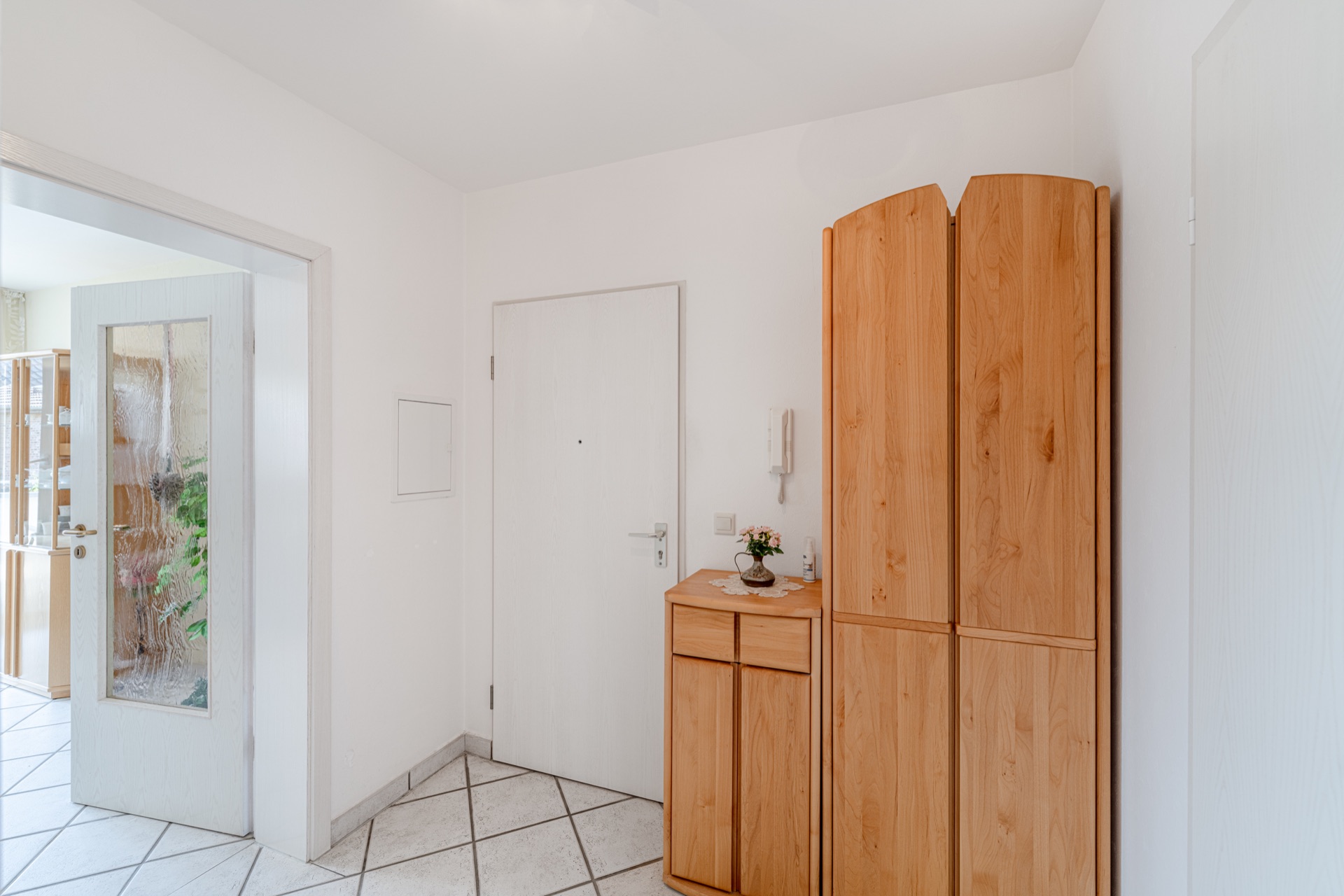 Heller Flur mit weißen Türen und Holzschränken, auf den Fliesenboden steht eine kleine Kommode mit Blumen.