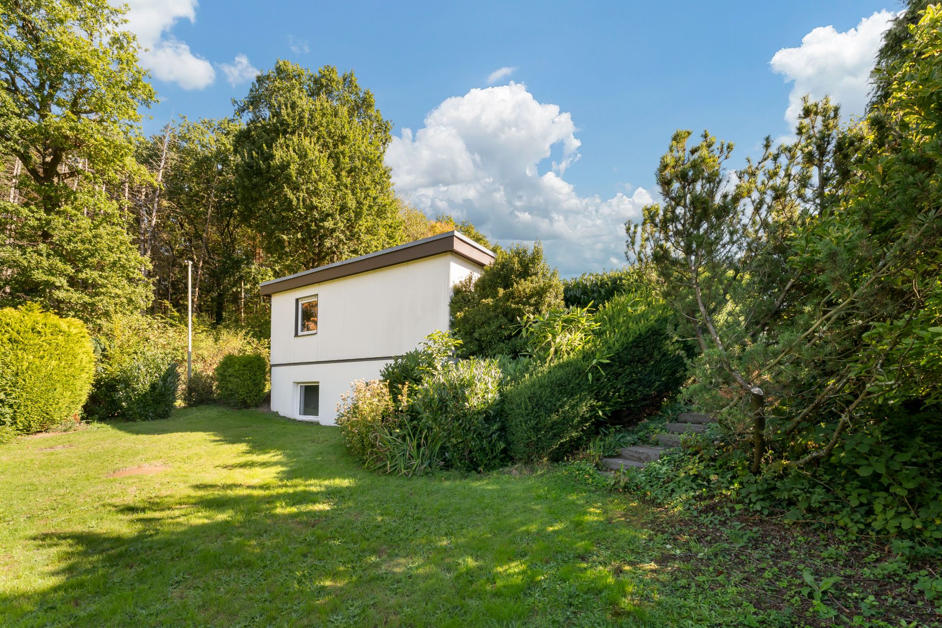 Kleines weißes Haus mit Flachdach im grünen Garten, umgeben von Bäumen und Büschen.