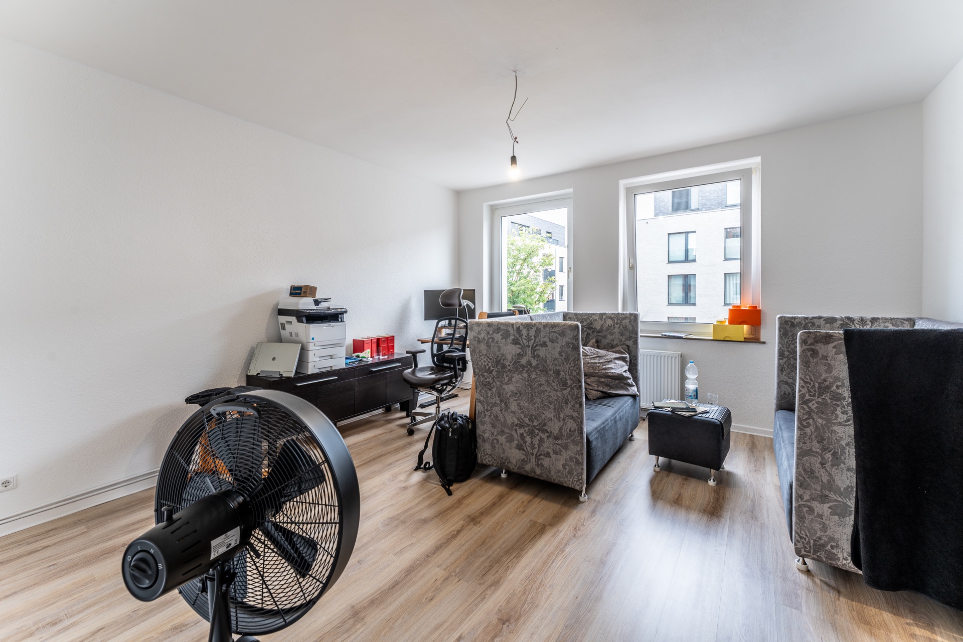 Helles Wohnzimmer mit Schreibtisch, Stuhl, grauen Sofas, Ventilator und zwei Fenstern.