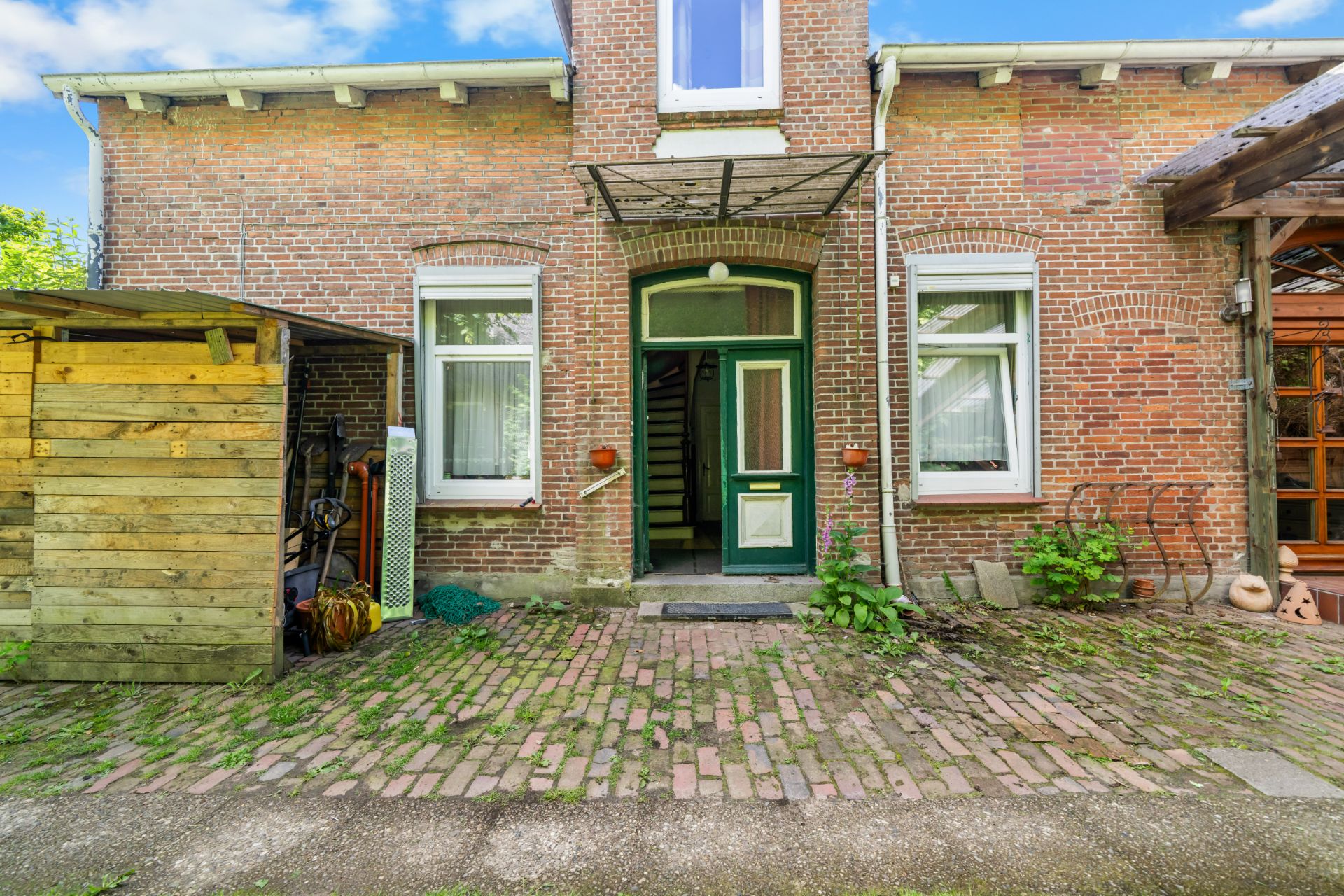Backsteinfassade eines Wohnhauses mit grüner Tür, zwei Fenstern und kleinem Holzschuppen neben dem Eingang.