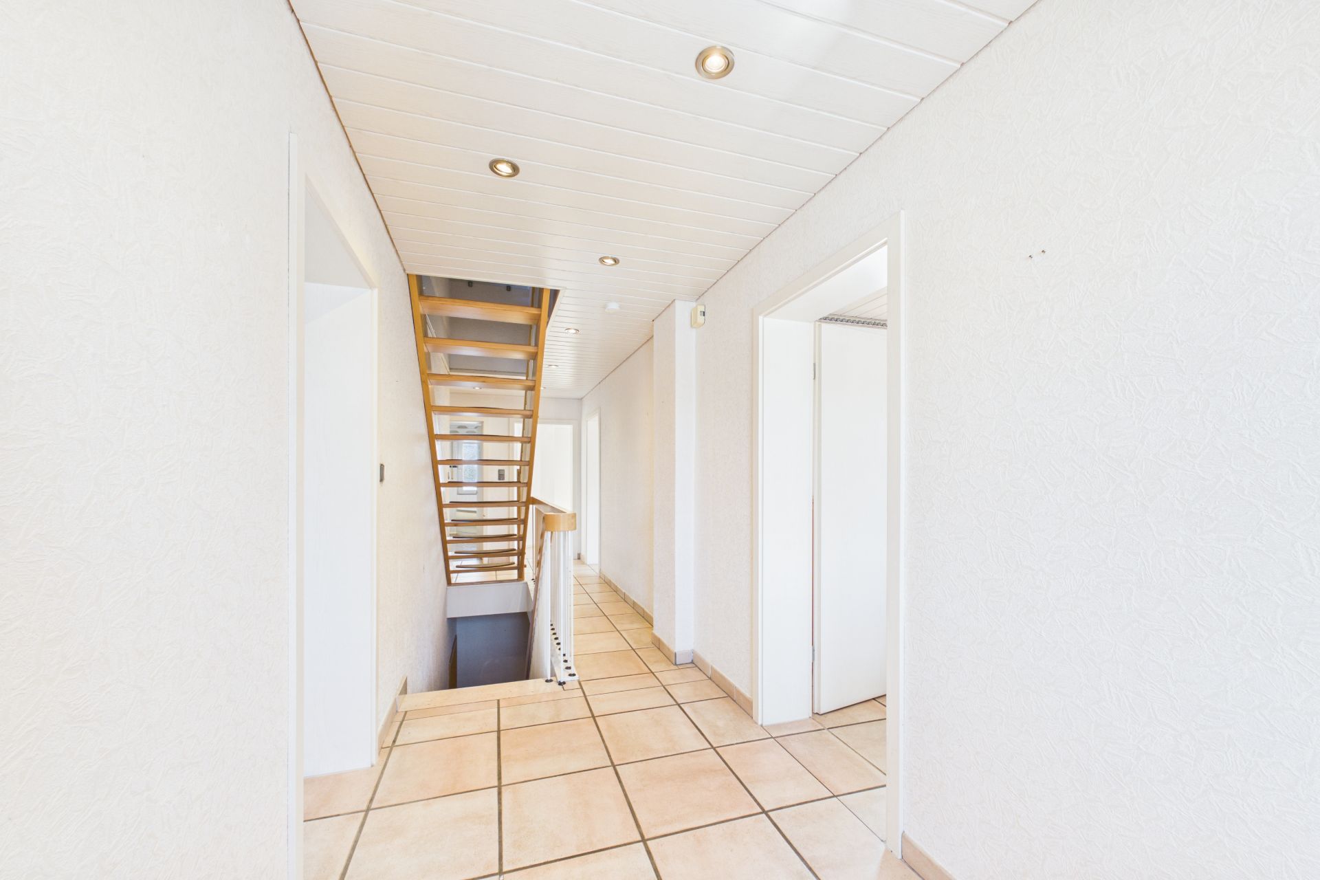 Heller Flur mit Fliesenboden, weißen Wänden, mehreren Türen und einer Holztreppe.