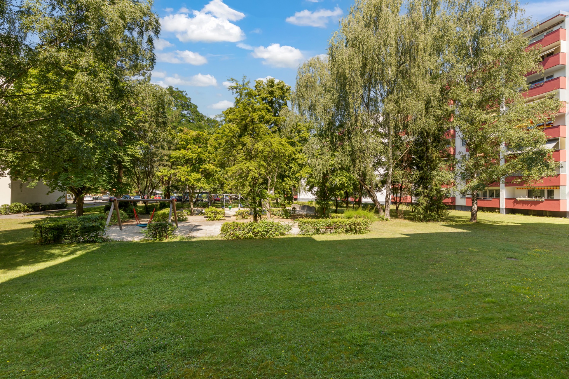Grüne Wiese mit Bäumen und Spielplatz, daneben ein mehrstöckiges Wohnhaus.
