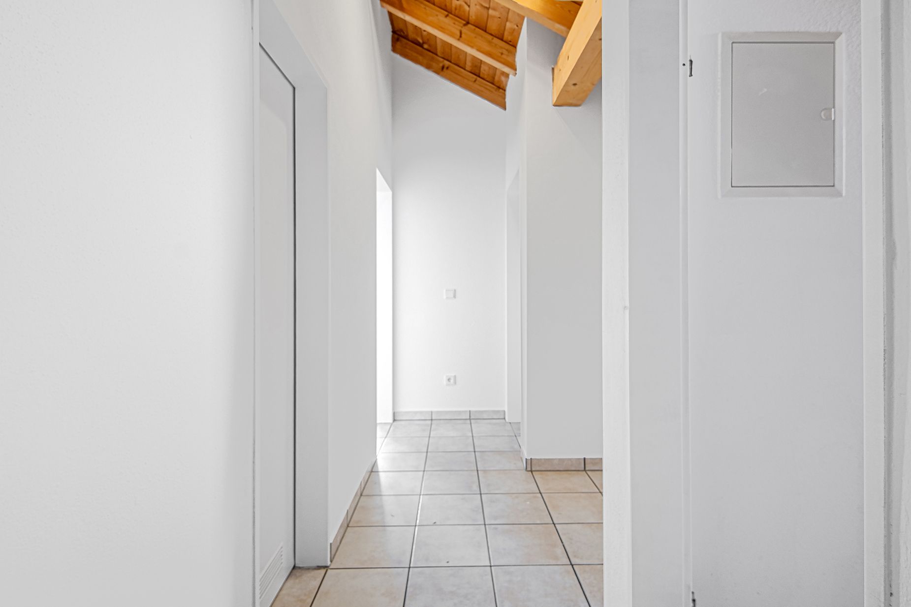 Heller Flur mit weißen Wänden, Fliesenboden und sichtbarer Holzdecke.