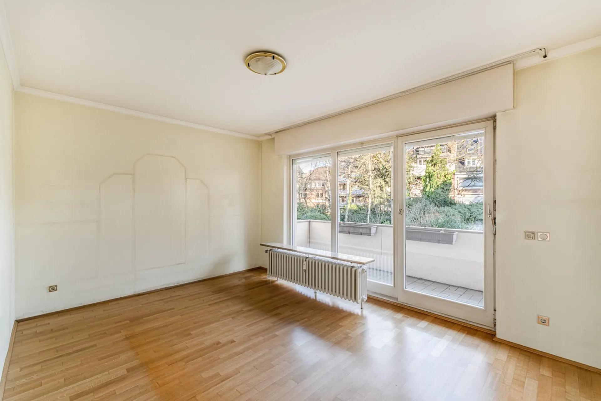 Helles, leeres Zimmer mit Parkettboden und großer Fensterfront mit Zugang zum Balkon.