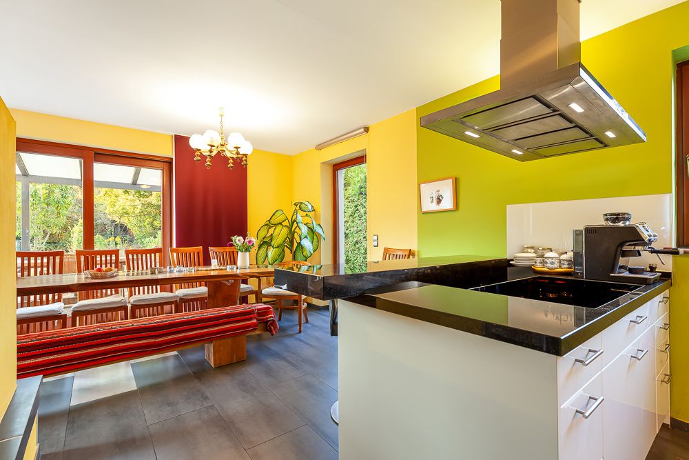 Helle Wohnküche mit langem Esstisch, Stühlen, Bank, Kochinsel und großen Fenstern.