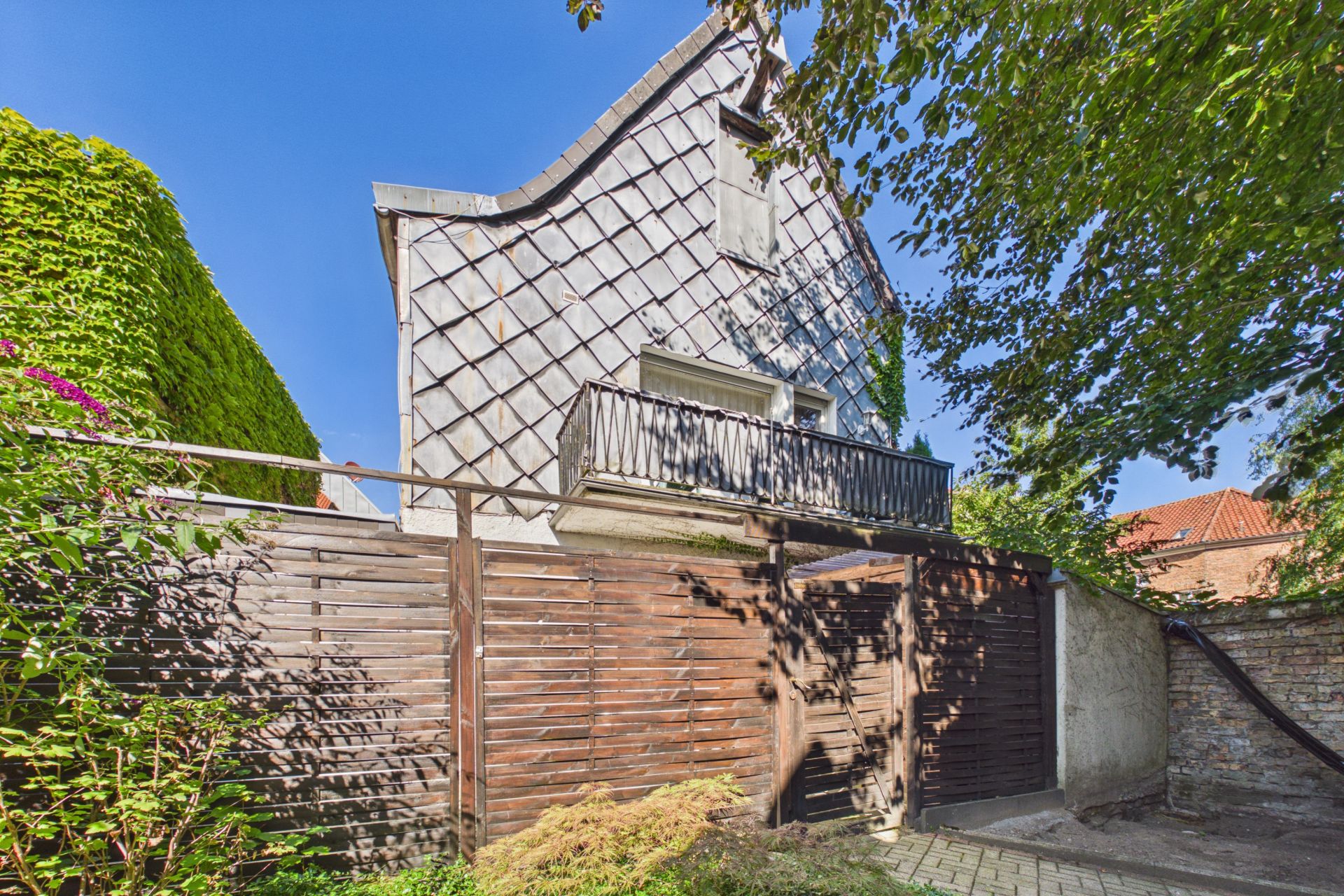Haus mit Holzlattenzaun, Balkon und Bäumen im Vordergrund.