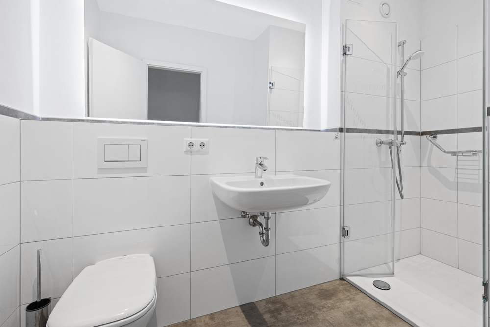 Modernes, weiß gefliestes Badezimmer mit WC, Waschbecken und offener Dusche.