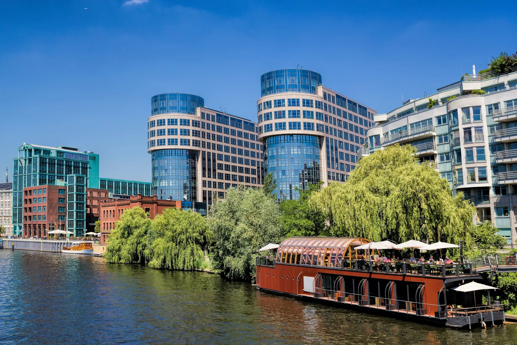 Moderne Wohngebäude und Bäume an einem Fluss mit einem Restaurantboot im Vordergrund.