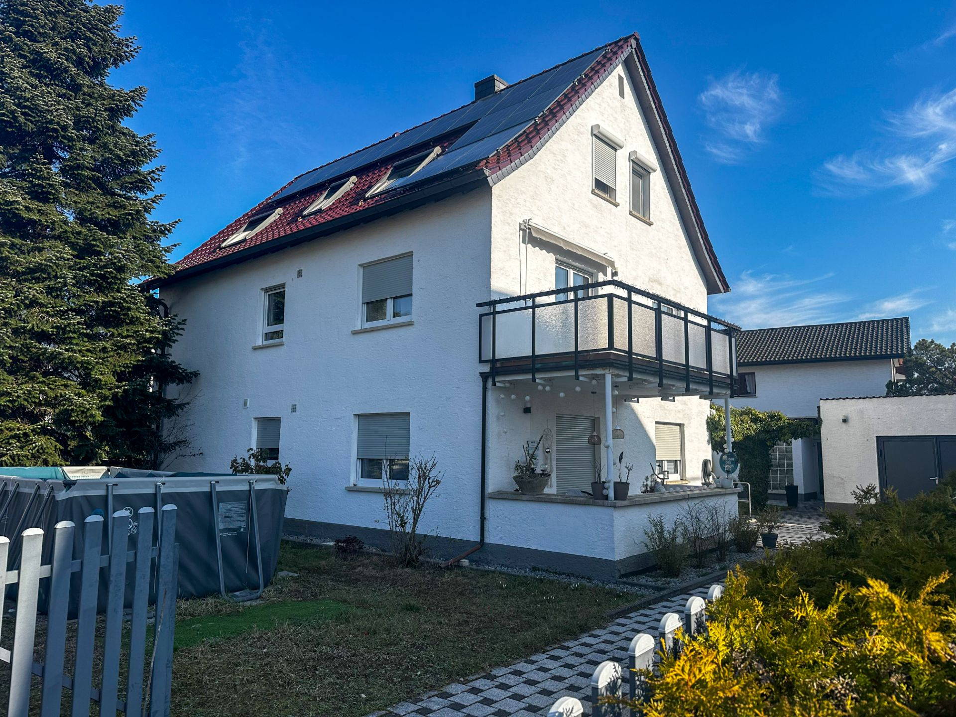 Weißes Einfamilienhaus mit Balkon, Garten und mehreren Fenstern an einem sonnigen Tag.