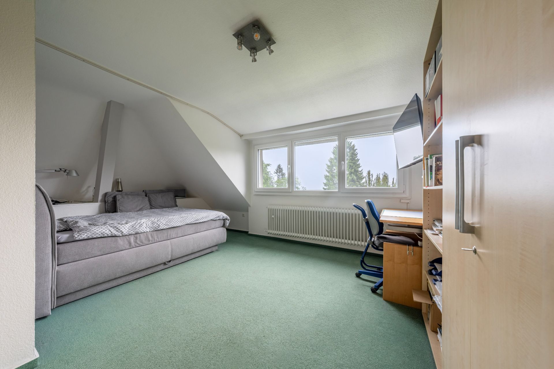Helles Zimmer mit Sofa, Schreibtisch, Regal und großem Fenster mit Blick ins Grüne.