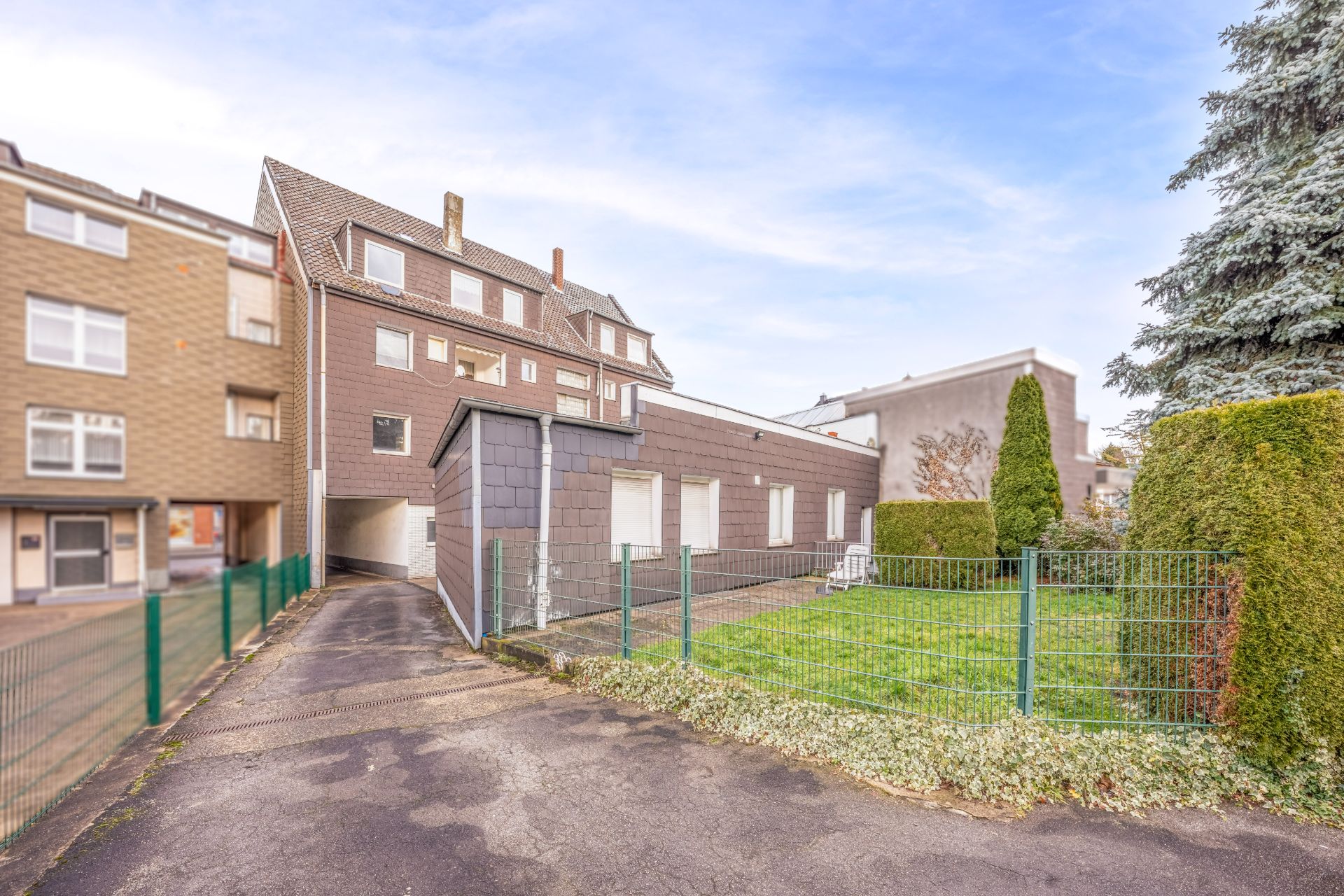 Mehrfamilienhaus mit Einfahrt, Vorgarten und umzäuntem Grünbereich, umgeben von weiteren Gebäuden.