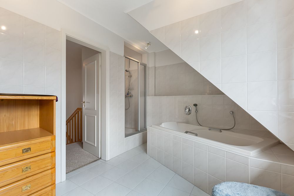 Helles Badezimmer mit Badewanne, Dusche, weißem Fliesenboden und Holzkommode.