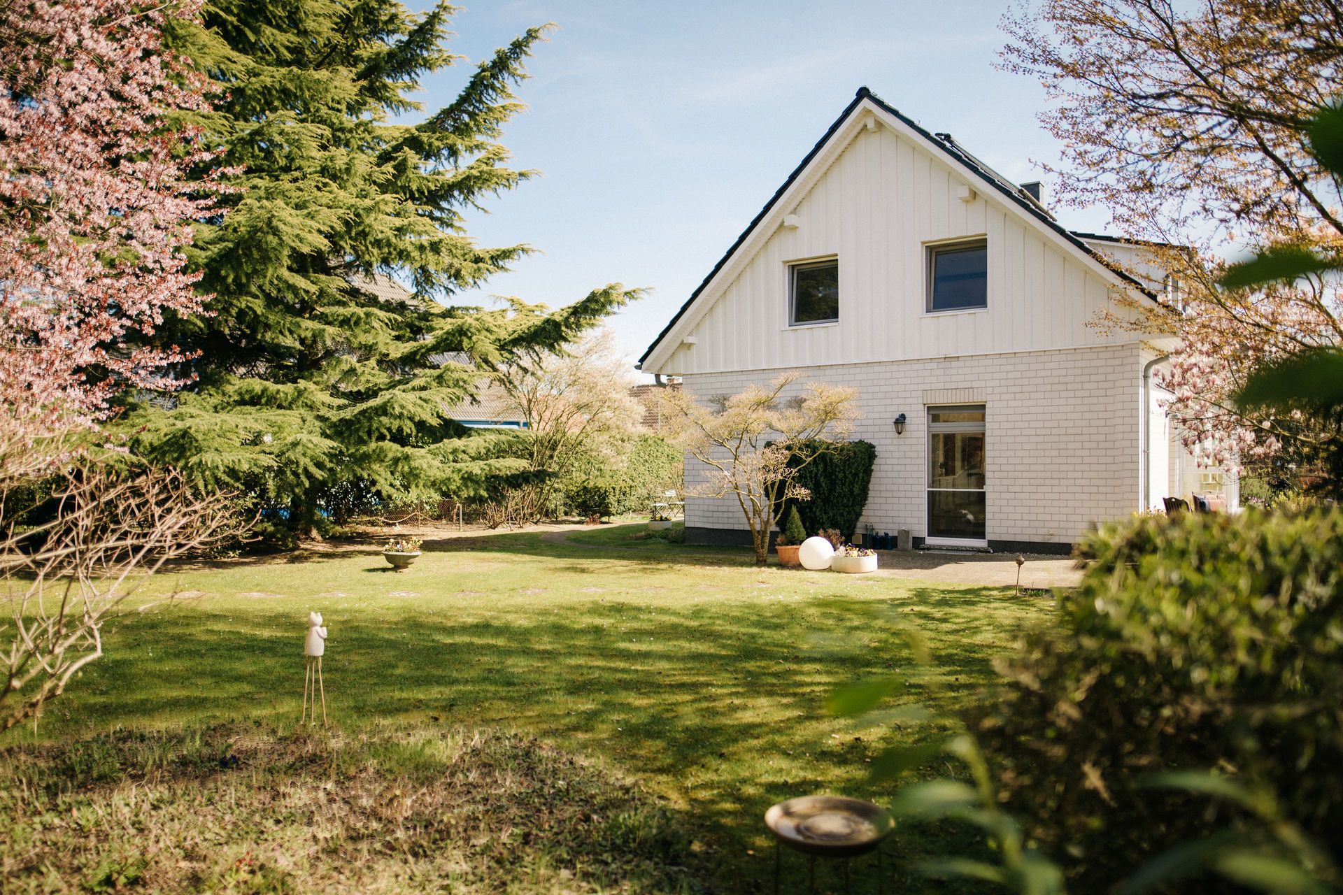 Weißes Einfamilienhaus mit Garten, Bäumen und Rasen an einem sonnigen Tag.