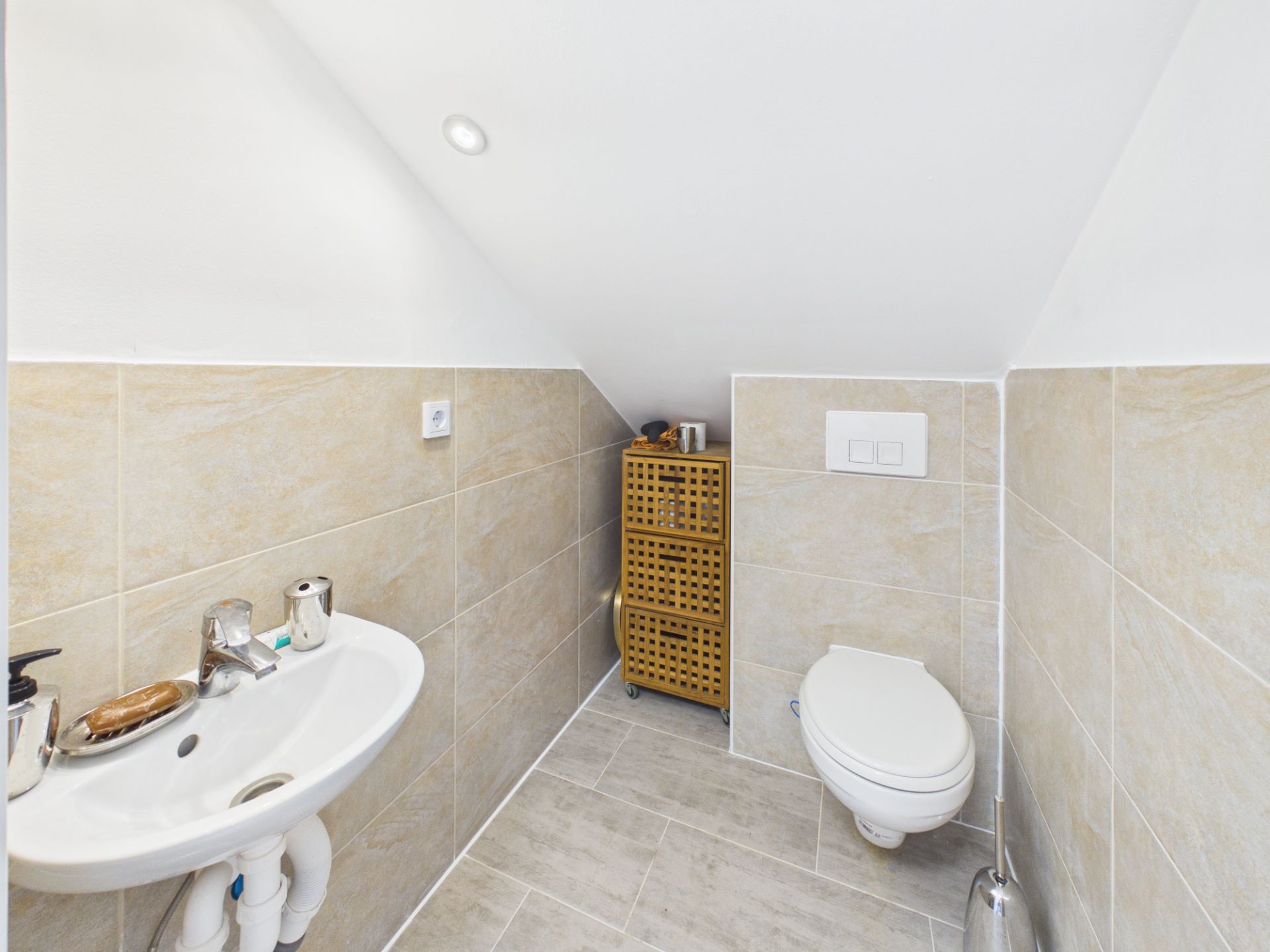 Kleines Bad mit Wandtoilette, Waschbecken und Schrank unter einer Dachschräge.