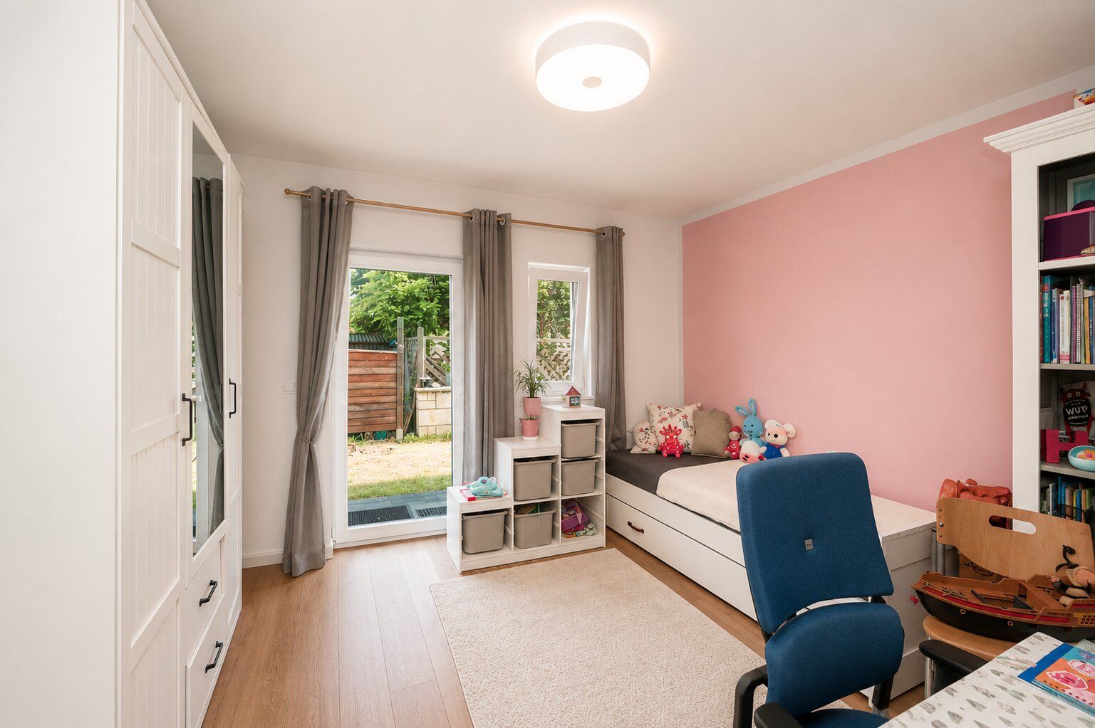 Helles Kinderzimmer mit rosa Wand, Bett, Schreibtisch, Regal und Blick in den Garten.