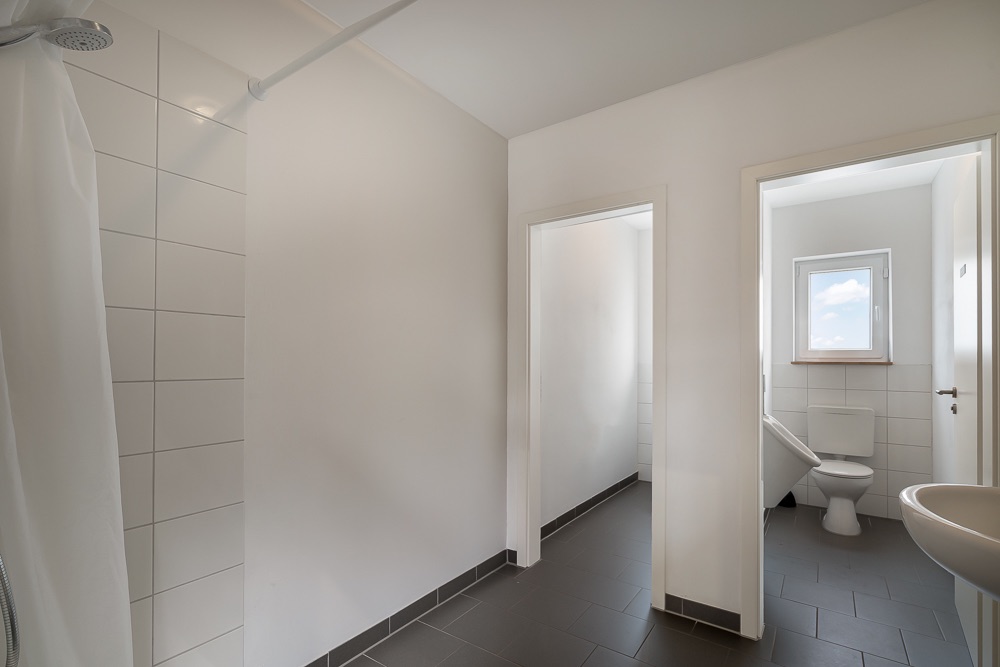 Helles Badezimmer mit Dusche, Waschbecken und angrenzendem Raum mit Toilette und Fenster.