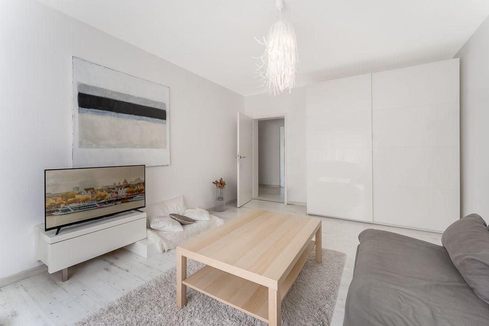 Helles Wohnzimmer mit grauem Sofa, Holztisch, Teppich, TV und großem weißen Schrank.