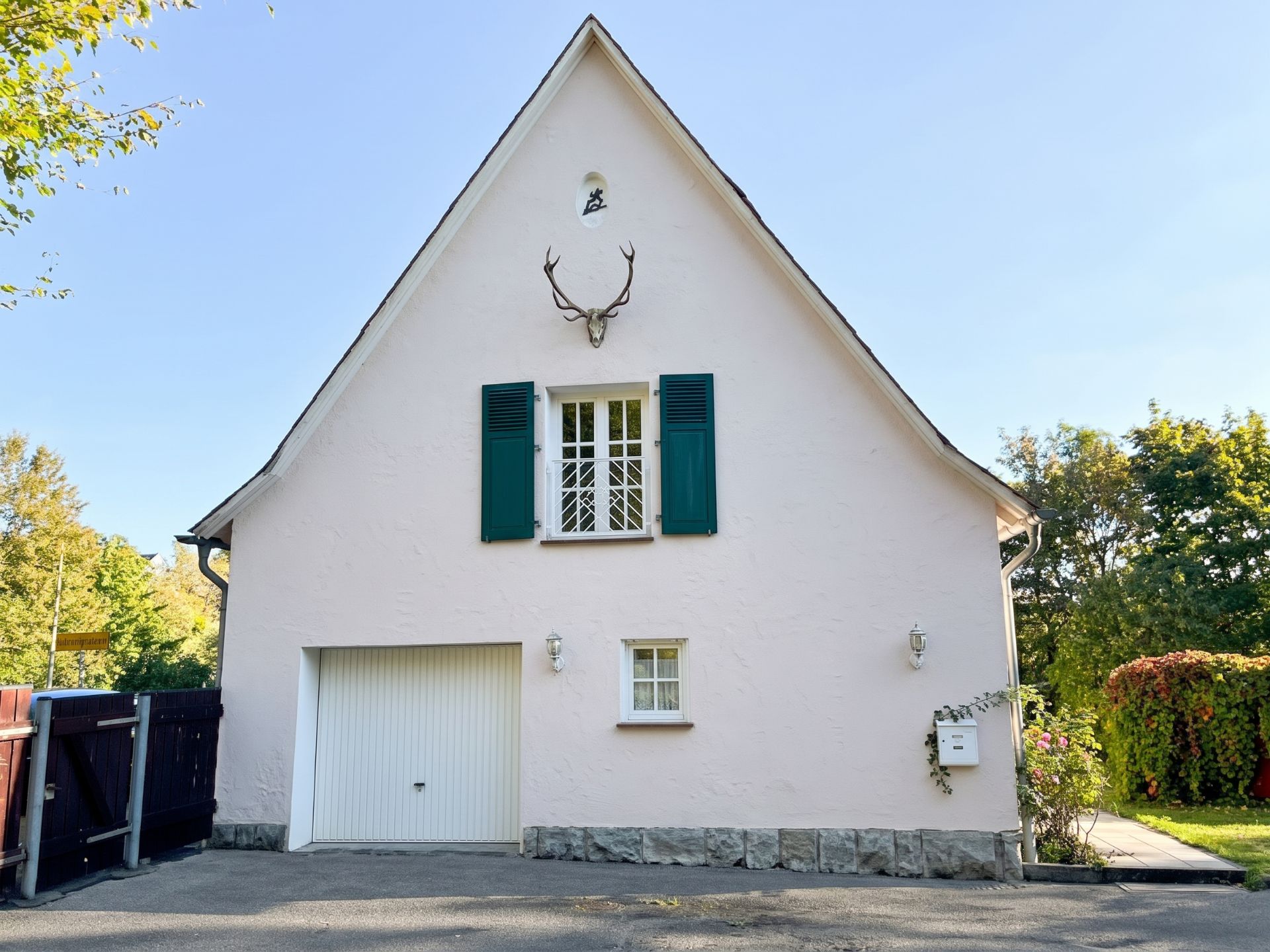 Weiße Hausfassade mit grünem Fensterladen, Garage und Hirschgeweih über dem Fenster.