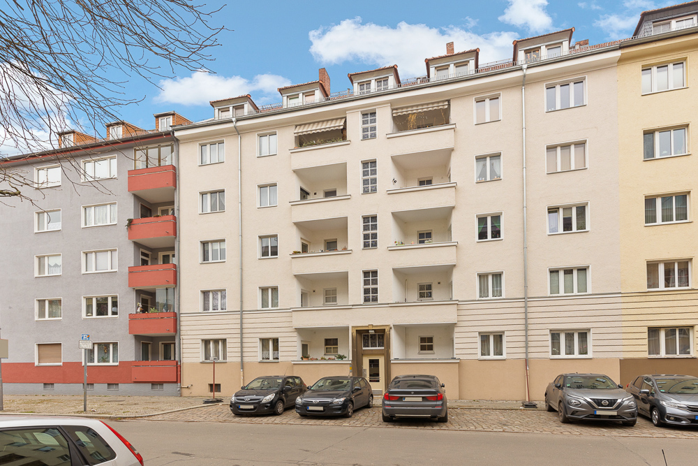 Mehrstöckiges Wohnhaus mit Balkonen, geparkten Autos davor und blauem Himmel im Hintergrund.